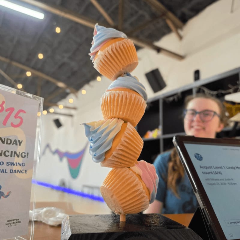 A stack of four cupcakes with blue and pink frosting stands in the foreground, while a smiling woman in glasses is visible in the blurred background.