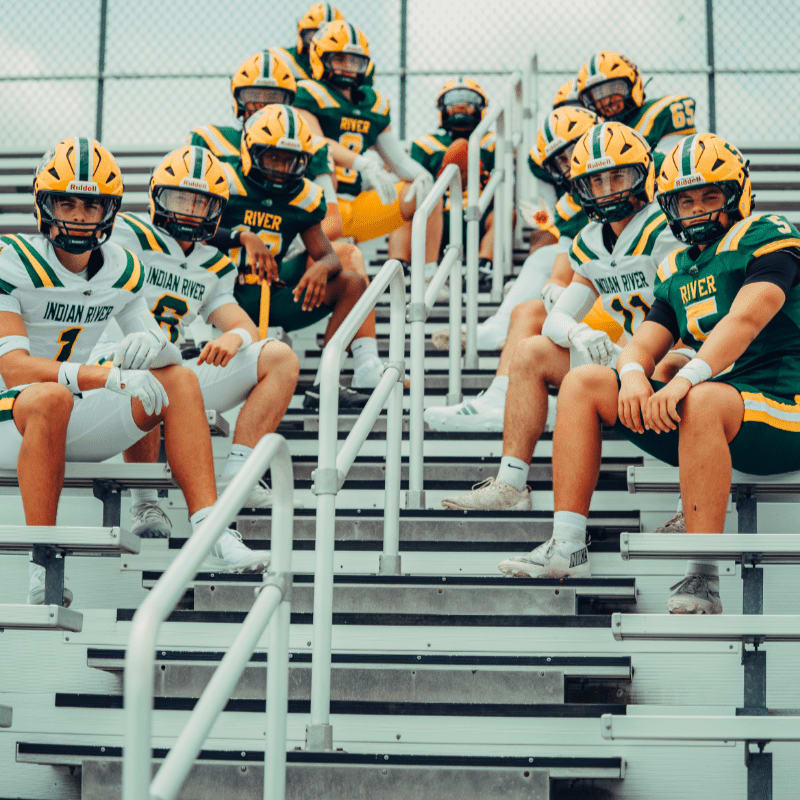 A group of young football players in green and yellow uniforms sit on bleachers in the foreground, with more players and a cloudy sky in the background.