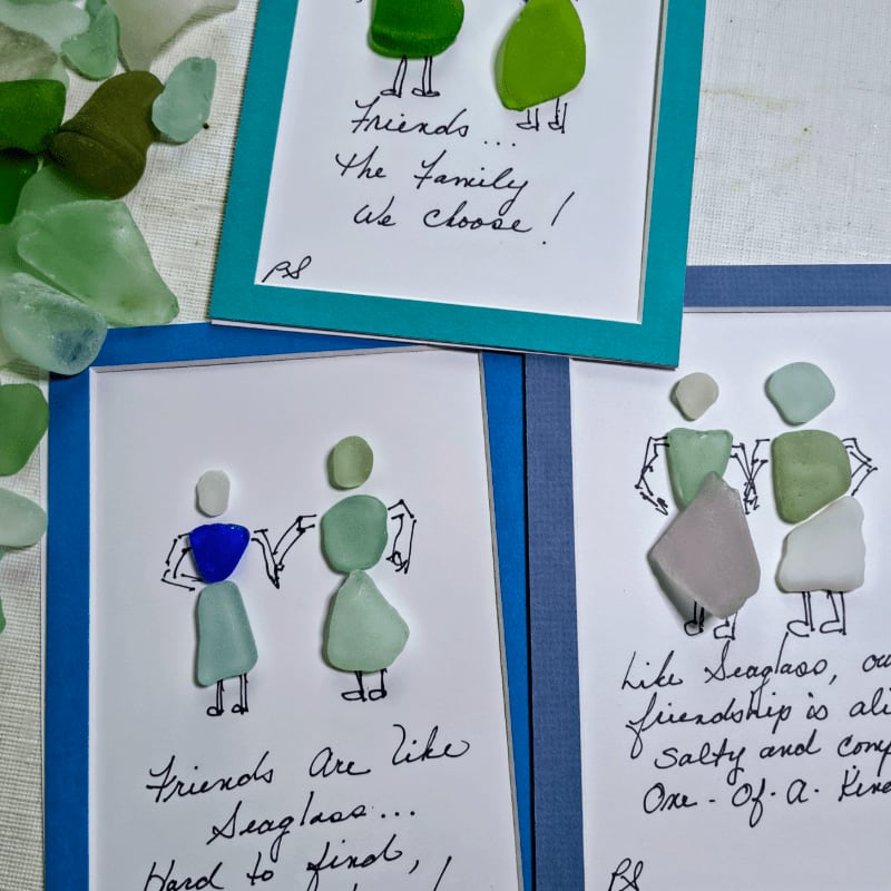 In the foreground, three framed artworks made of sea glass figures and handwritten quotes are arranged, while the background is scattered with various pieces of sea glass.