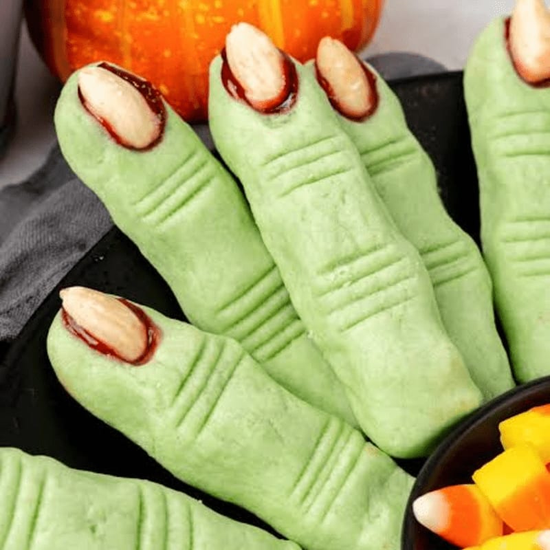 Green witch finger cookies with almond fingernails are arranged on a black plate in front of a pumpkin and a bowl of candy corn.