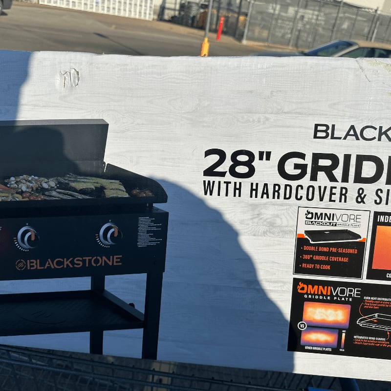A Blackstone 28" griddle with a hardcover sits in the foreground, with a parking lot and buildings in the background.