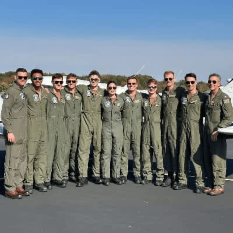 A group of ten pilots in green flight suits stand on an airfield in front of a small airplane under a clear blue sky.
