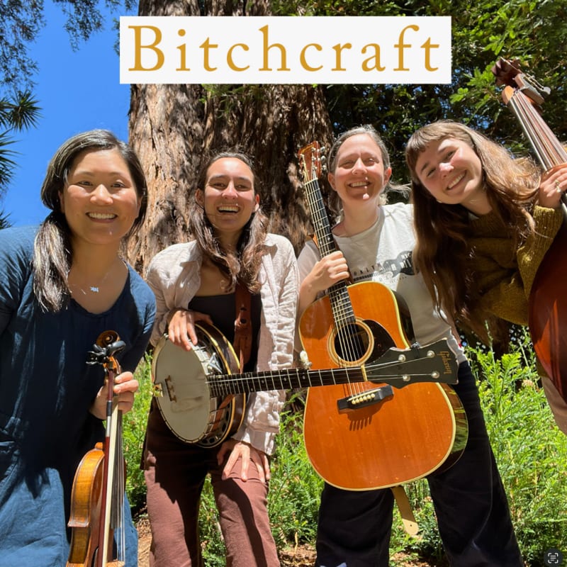 Four smiling women, holding musical instruments, stand in front of a large tree and lush greenery.