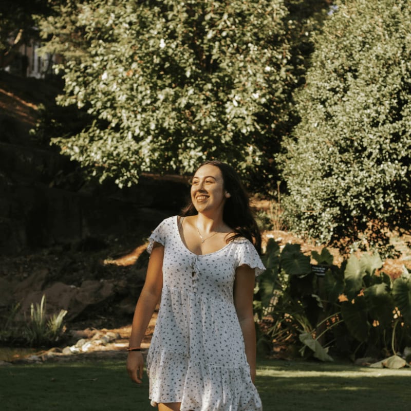 A young woman in a white polka-dot dress smiles as she walks across a grassy lawn with lush green trees in the background.