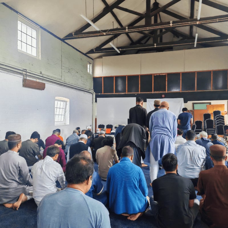 Men in prayer rugs fill the foreground of a large room with a high ceiling and windows in the background.