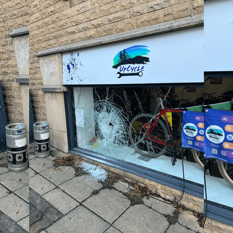 Shattered glass from a broken storefront window reveals bicycles inside, with two metal kegs standing on the sidewalk in the foreground.