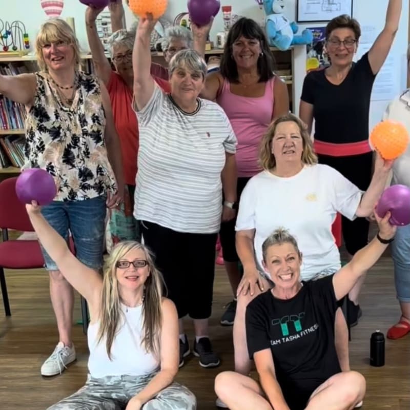 A group of women of various ages are happily exercising with colorful balls in a room with bookshelves in the background.