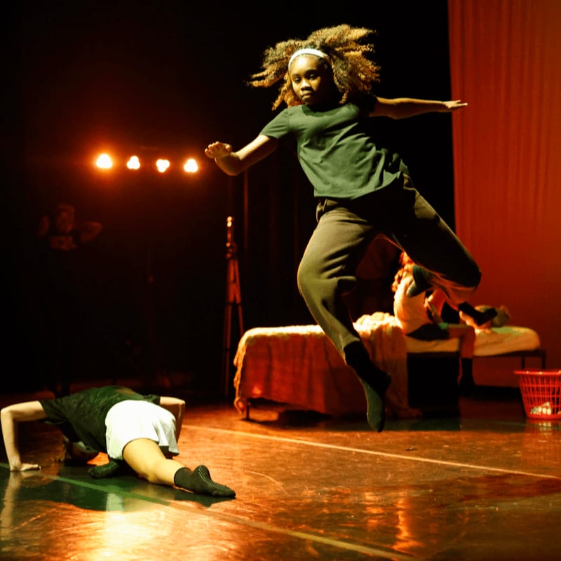A dancer leaps in the air on a stage with another dancer on the floor in the foreground, while a bed and red curtains form the background.