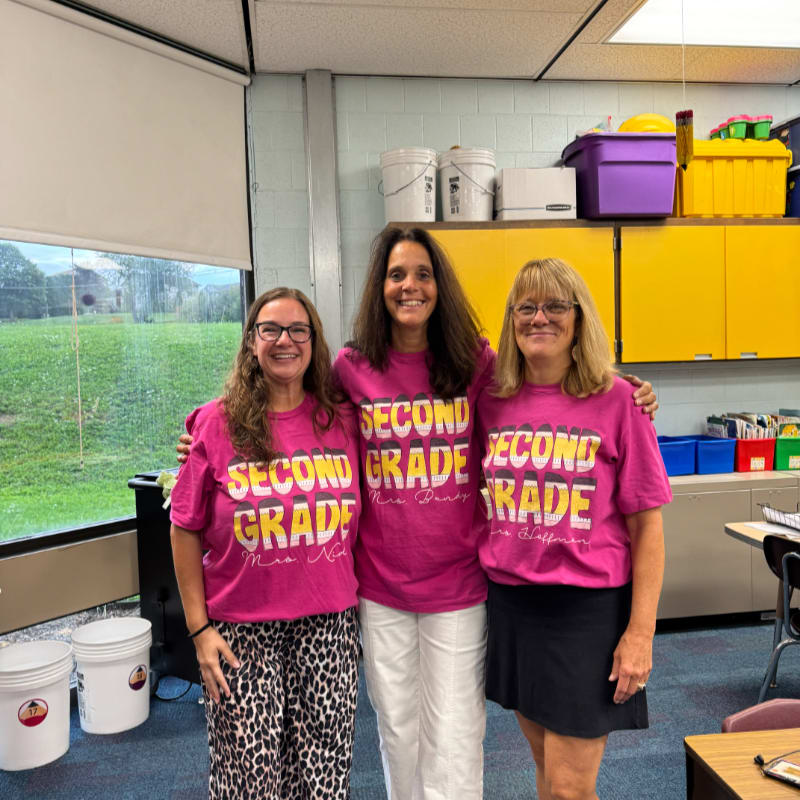 Three women in matching "Second Grade" t-shirts stand in a classroom with shelves of supplies and a window showing greenery in the background.