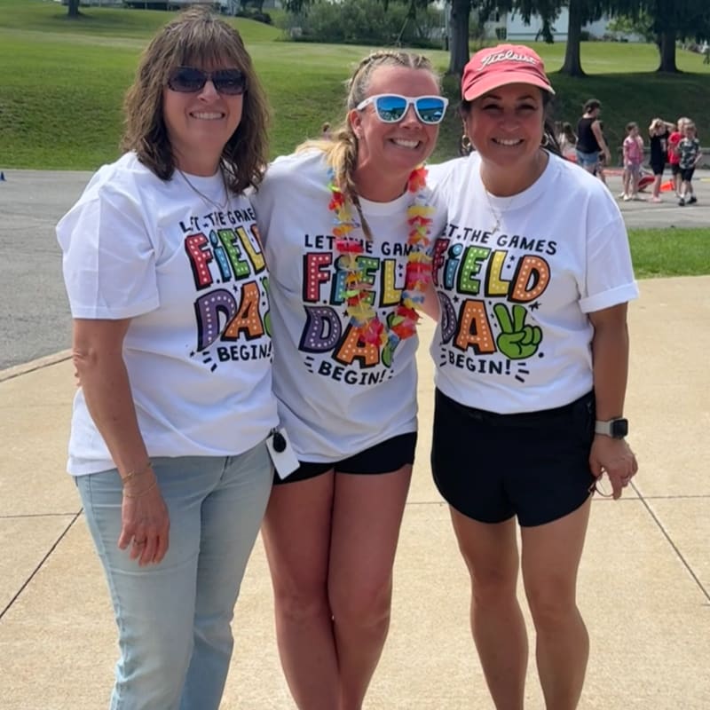 Three smiling women in "Field Day" t-shirts pose in the foreground with a grassy field and people in the background.