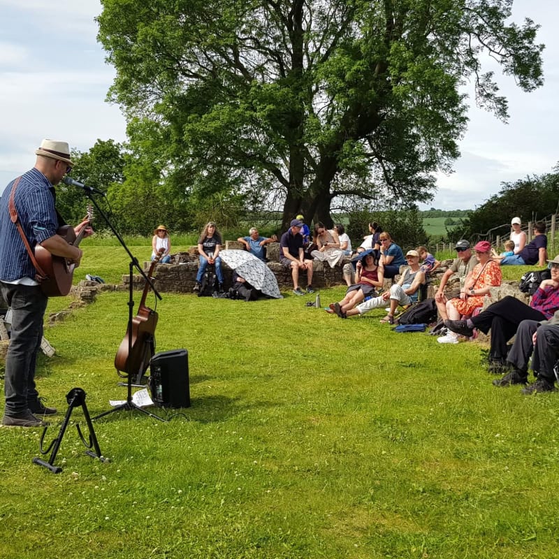 A man in a hat plays guitar for an audience seated on a grassy hill, with a large tree and distant fields in the background.