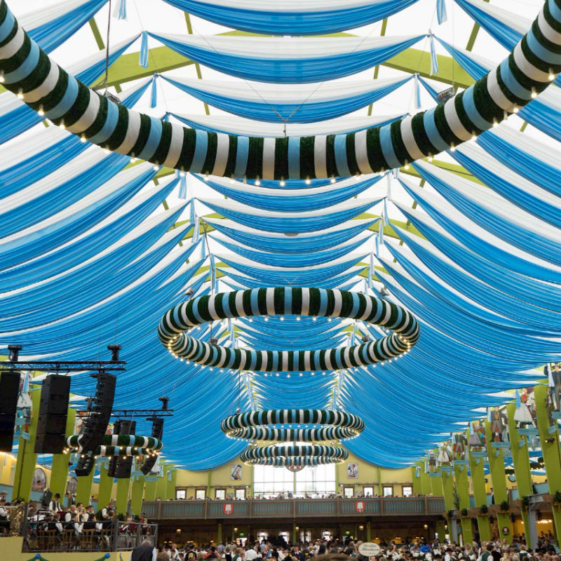 Blue and white fabric drapes form a tented ceiling above a lively beer hall filled with people and decorated with striped rings.