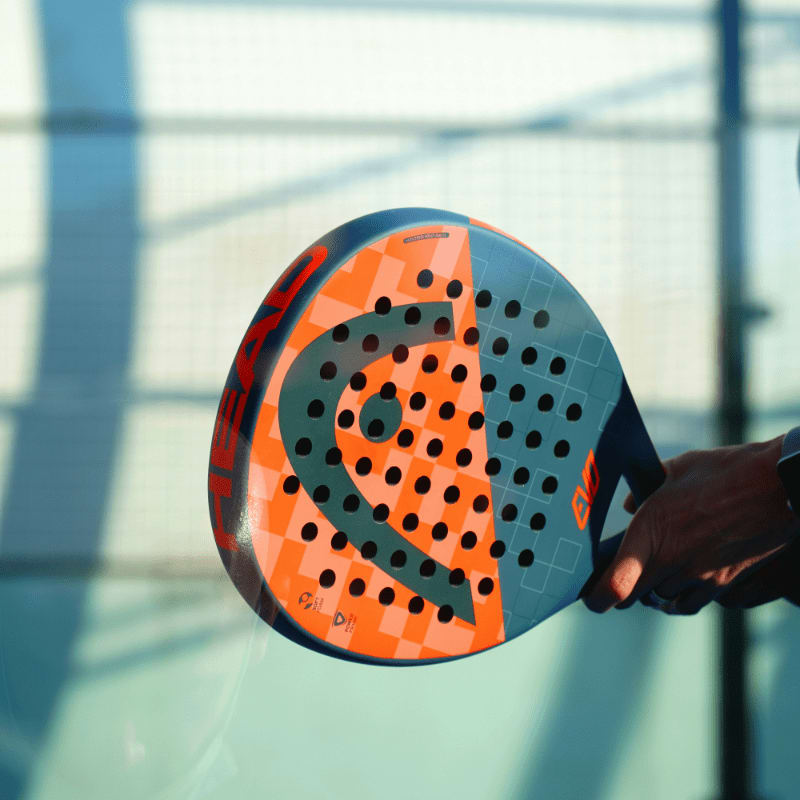 A person holding an orange and blue padel racket on a court
