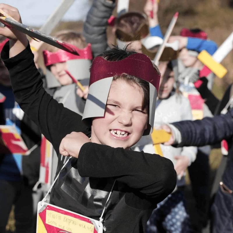 A young boy in a cardboard crown and armor strikes a pose with a toy sword in the foreground, while other children in similar costumes are visible in the blurred background.