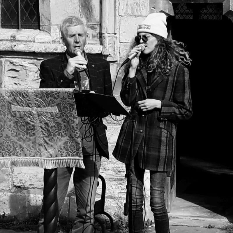 A man and a young woman sing into microphones in front of a stone building.