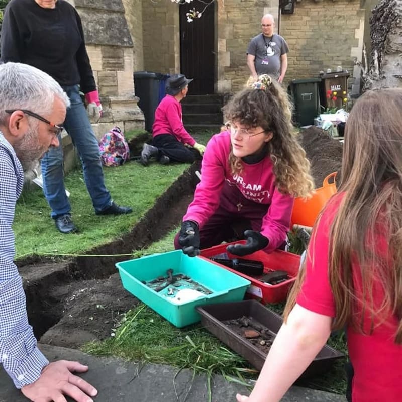 In the foreground, people are sifting through dirt in shallow trays, while in the background, others are working in a garden area near a building.