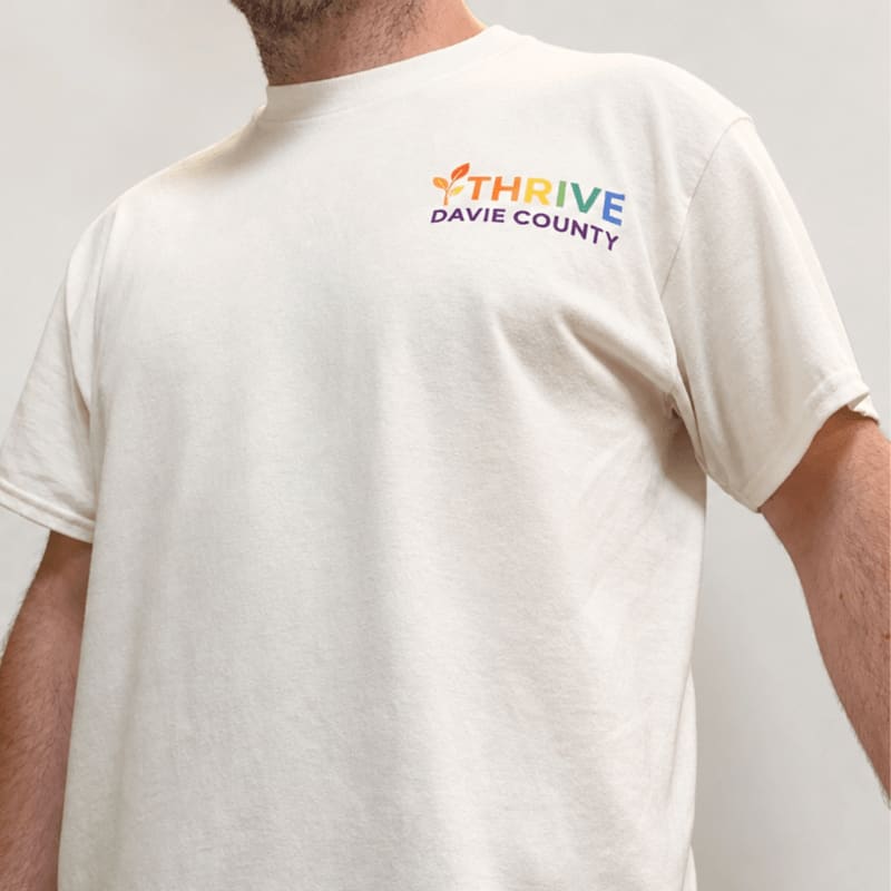 A person wears a cream-colored t-shirt with a colorful "THRIVE DAVIE COUNTY" logo on the upper left chest, set against a plain, light-colored background.