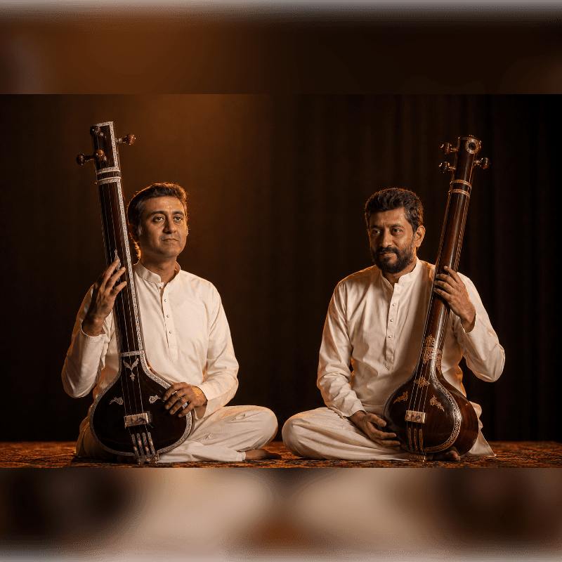Two men in white tunics sit cross-legged on a patterned rug, each holding a tanpura, with a dark, draped background.