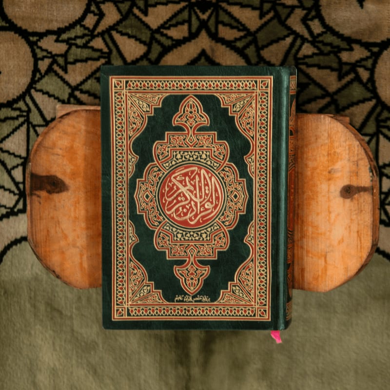 A green Quran on a wooden book stand over a patterned prayer rug