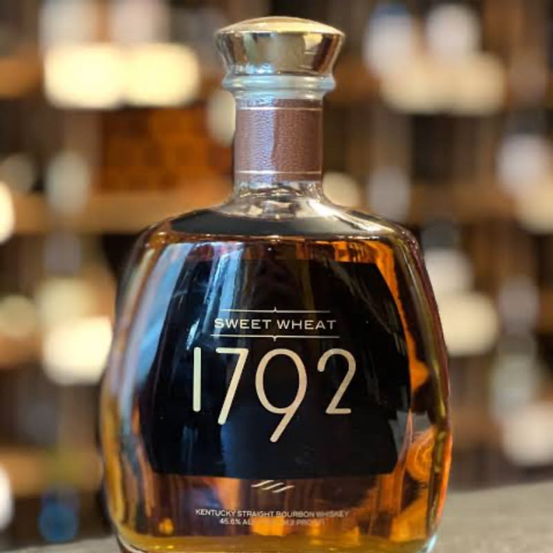 A bottle of 1792 Sweet Wheat Kentucky Straight Bourbon Whiskey stands in the foreground, with shelves of other bottles blurred in the background.
