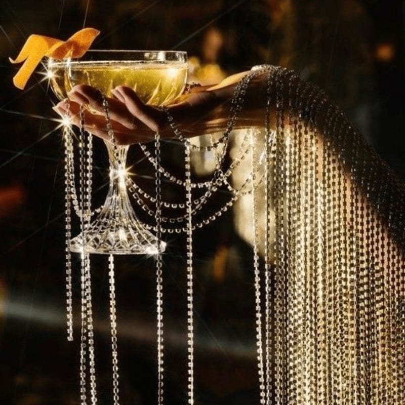 A hand adorned with sparkling rhinestones holds a champagne coupe with an orange peel garnish, set against a softly blurred, warm-toned background.