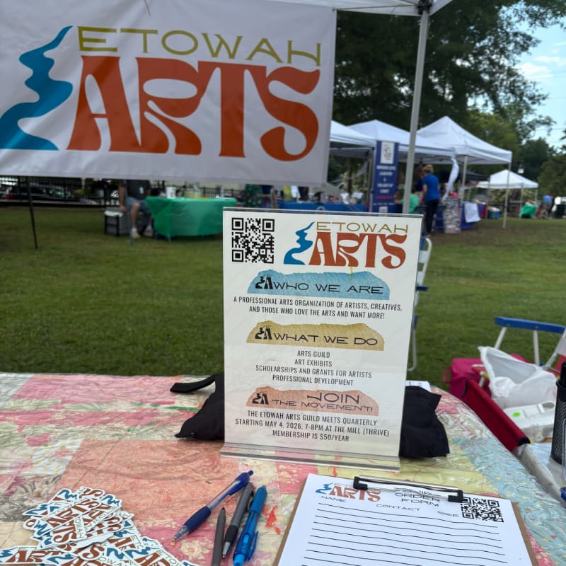 A table in the foreground displays pens, stickers, and a clipboard with information about Etowah Arts, while a banner and tents are visible in the blurred background of an outdoor event.