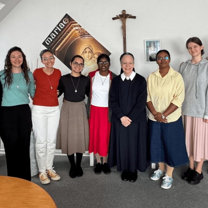 Seven women, some in religious habits, stand in a room with a crucifix and religious artwork on the wall, while a wooden table is visible in the foreground.