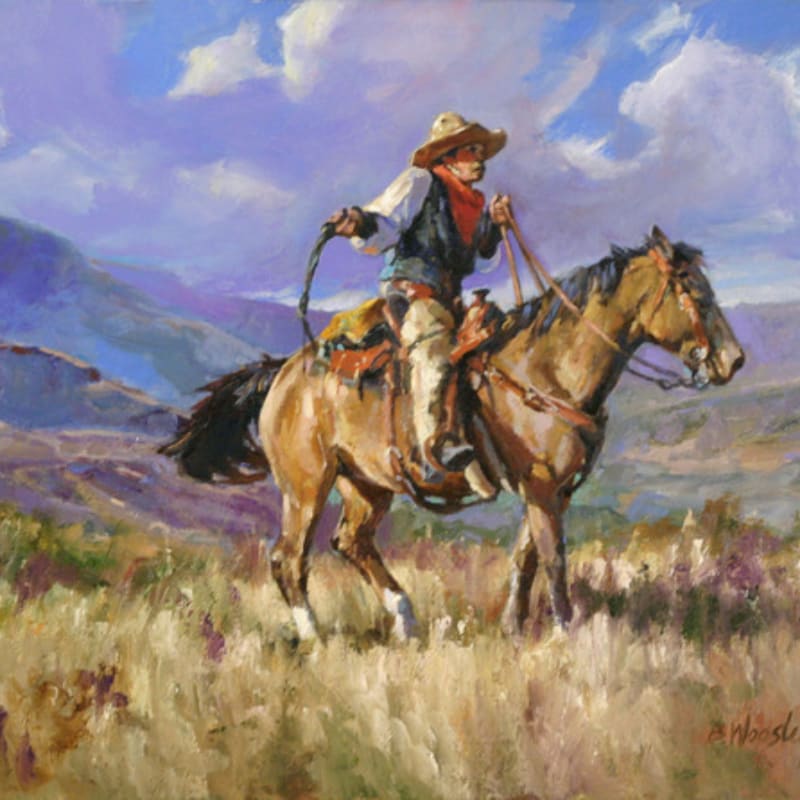 A cowboy on horseback rides through a field of dry grass with purple mountains and a cloudy sky in the background.