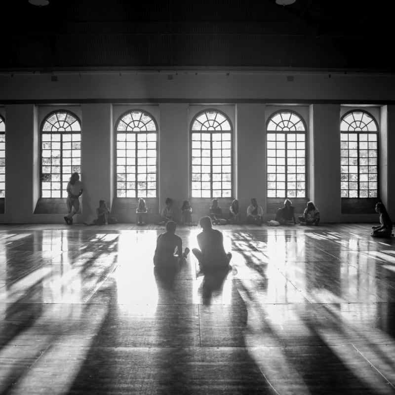 Silhouetted figures sit in meditation on a sunlit wooden floor, with arched windows casting striped shadows across the room.