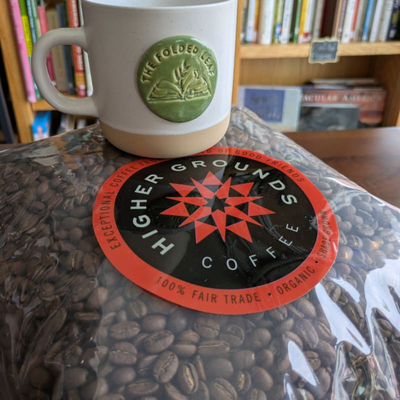 A white mug with a green emblem sits atop a bag of coffee beans, with bookshelves in the background.