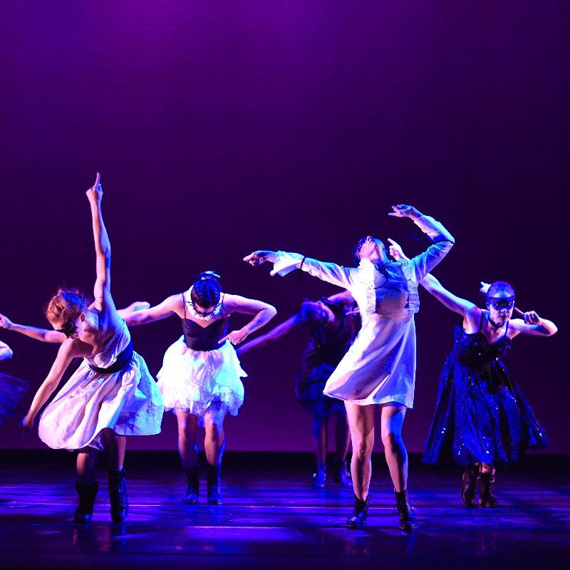 Dancers in white and dark costumes perform on a stage with a purple backdrop.