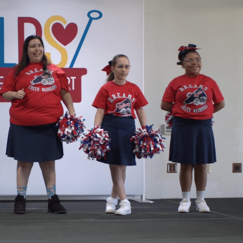 Three young women in red shirts and blue skirts, holding pom-poms, stand on a stage in front of a white backdrop with a colorful logo.