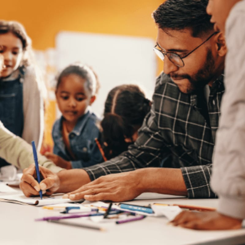 A man with glasses and a plaid shirt draws with a blue pen at a table with children, while a blurred orange background suggests a classroom.