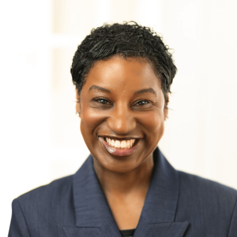 A smiling woman with short, dark hair wears a navy blazer in the foreground, against a softly blurred, light-colored background.