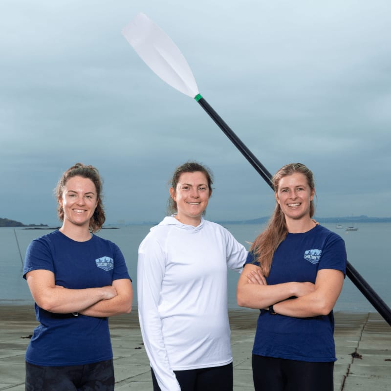 Three women in athletic wear stand with their arms crossed in the foreground, while a large oar is held diagonally behind them against a backdrop of a calm body of water with boats and a cloudy sky.