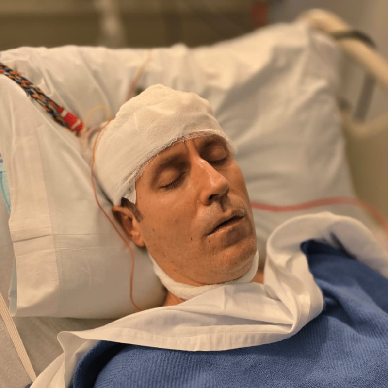 A man with a bandaged head rests in a hospital bed, with medical equipment visible in the blurred background.