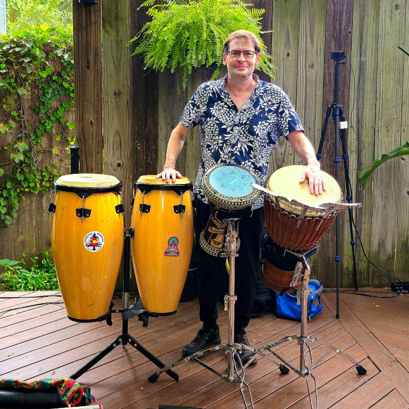 A man stands behind a collection of drums on a wooden deck, with lush greenery and a fence in the background.