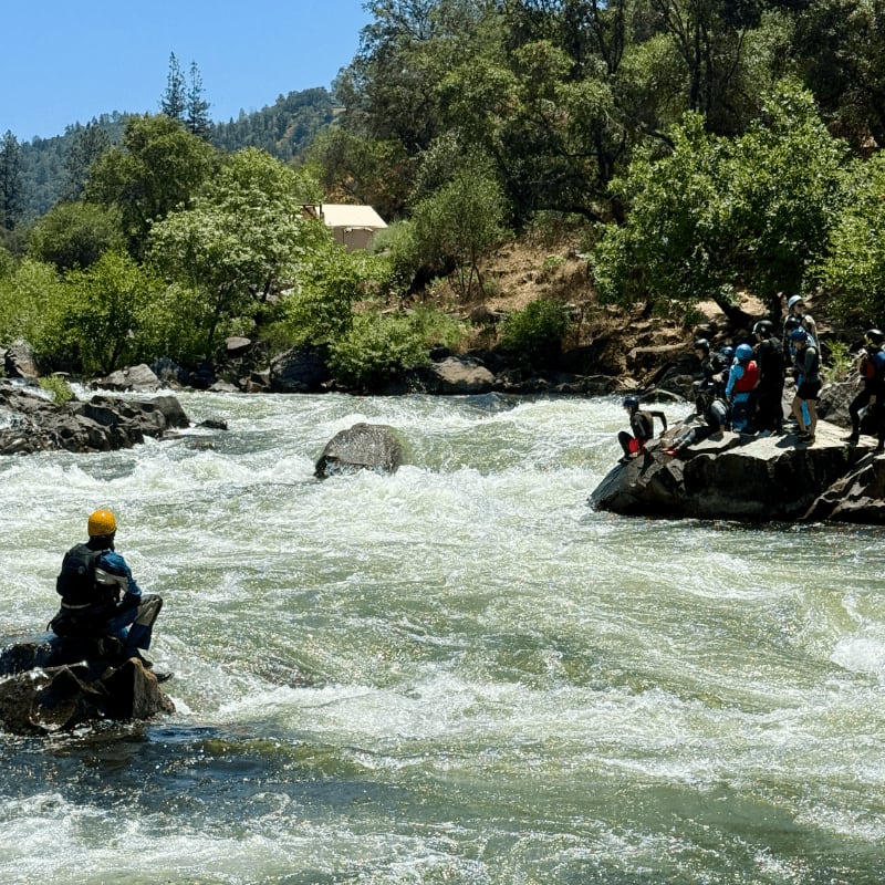 A lone kayaker in a yellow helmet sits on a rock in the foreground of a rushing river, while a group of people in colorful life vests stand on rocks in the background, surrounded by lush green trees and a clear blue sky.