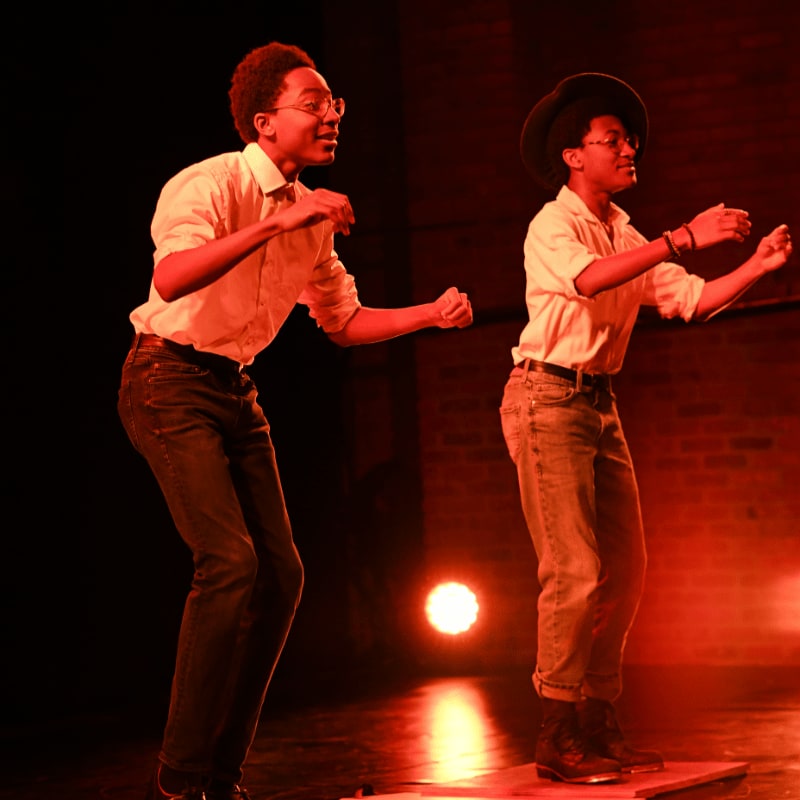 Two young men in light-colored shirts and jeans perform on a stage with a red-lit background and a single spotlight.