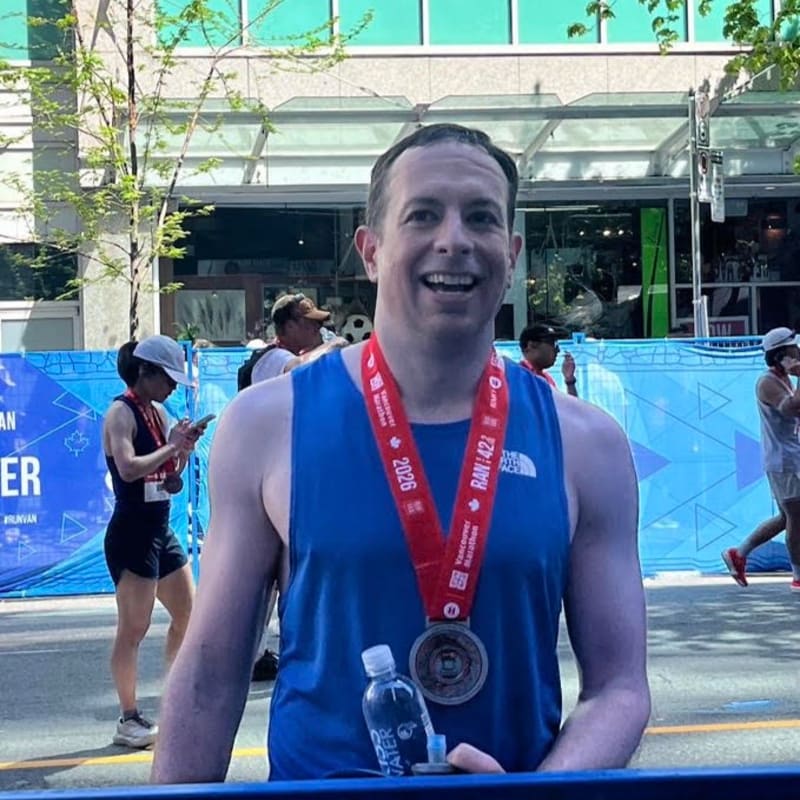 A smiling man in the foreground wears a medal around his neck, while in the background, other runners and spectators are visible near a blue banner.