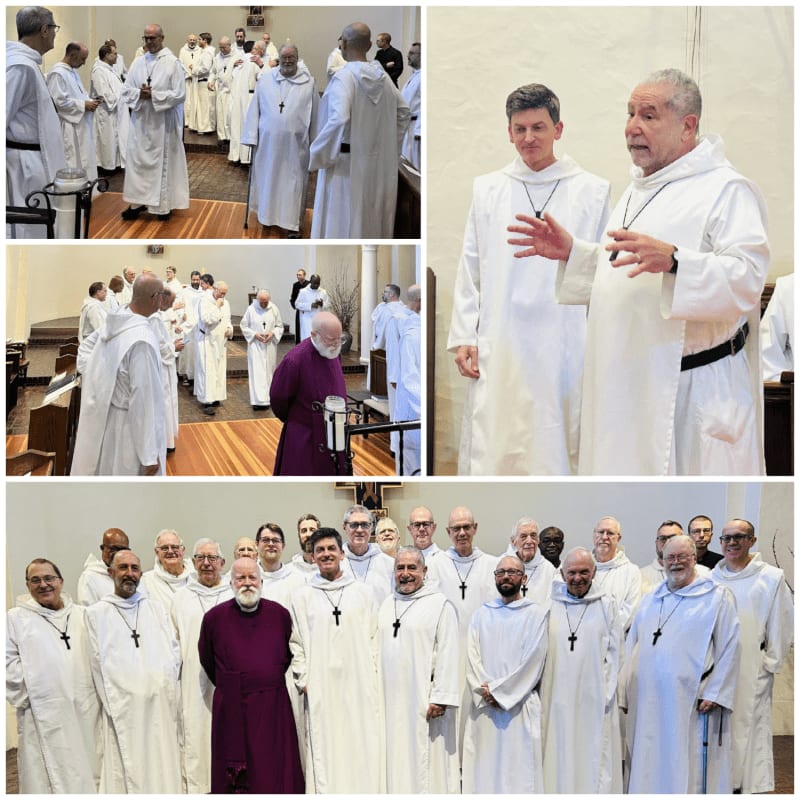 A group of men in white robes stand in a room with wooden floors and walls, while in the background, two men in white robes speak to each other.