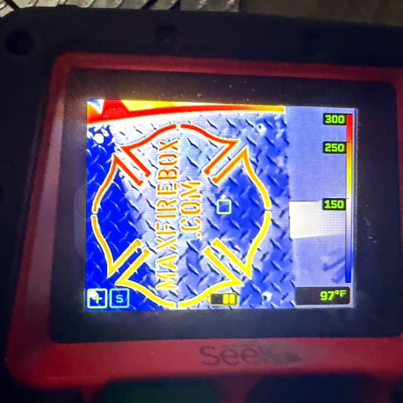 A thermal imaging camera displays a red and blue Maltese cross logo with "MAXFIREBOX.COM" written on it against a diamond plate background, with a temperature scale on the right.