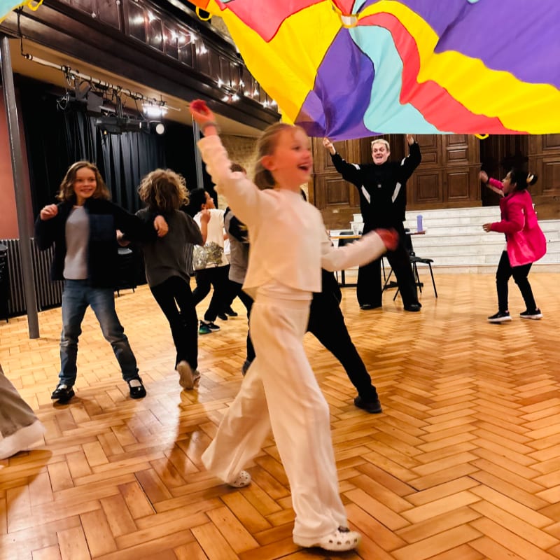 A young girl in white pants and top dances joyfully under a colorful parachute held by children and an adult in a room with a wooden floor and stage in the background.