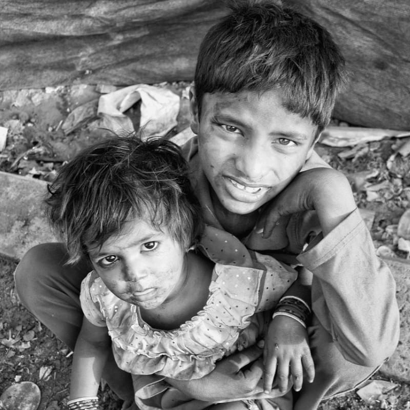 A young boy and girl, their faces smudged with dirt, sit close together in the foreground, with a rough, makeshift shelter visible in the background.