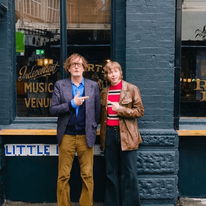 Two men in vintage clothing stand in front of a dark brick building with a sign for an independent music venue.
