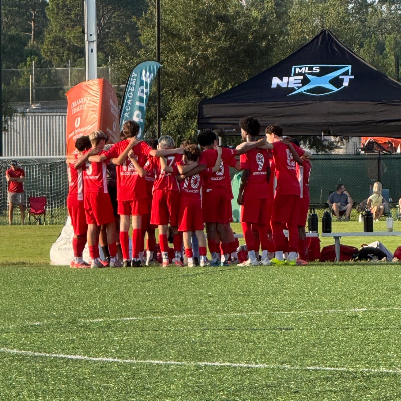 A group of young soccer players in red uniforms huddle together on a green field, with a tent and spectators in the background.