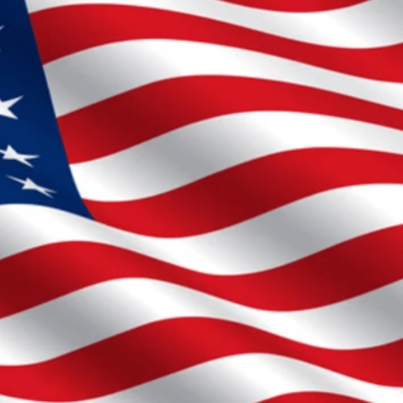 A close-up of the American flag fills the frame, with the blue union and white stars visible on the left and red and white stripes rippling across the rest of the image.