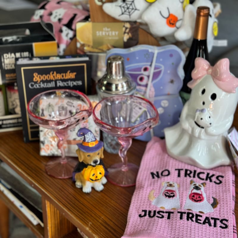 A Halloween-themed cocktail set with a dog figurine in the foreground sits on a wooden surface, with ghost decorations and a bottle of wine in the background.