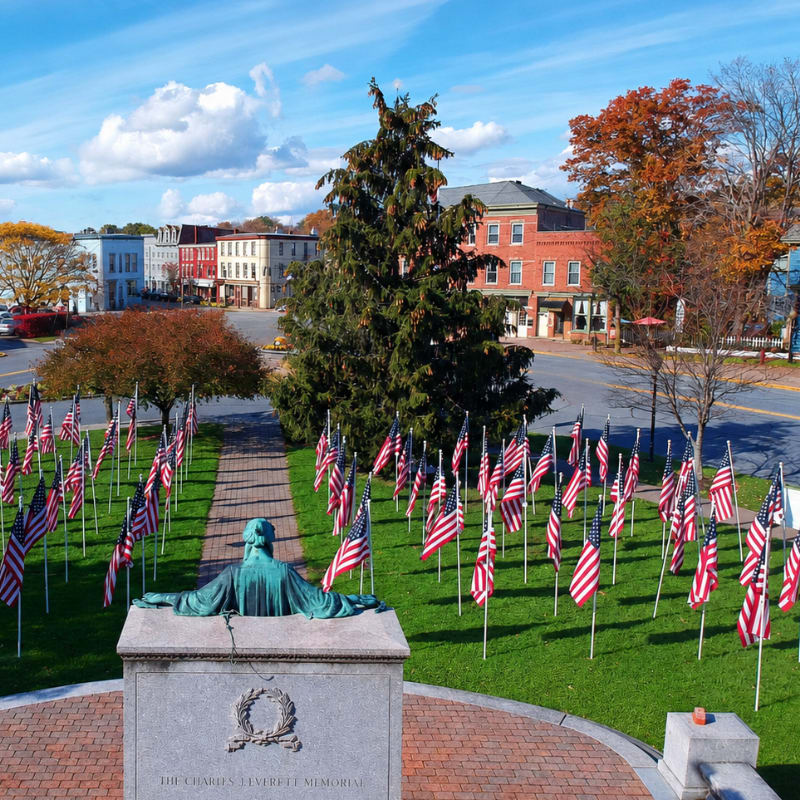 A statue of a man in a green patina overlooks a field of American flags in the foreground, with a quaint town and autumn foliage in the background under a bright blue sky.