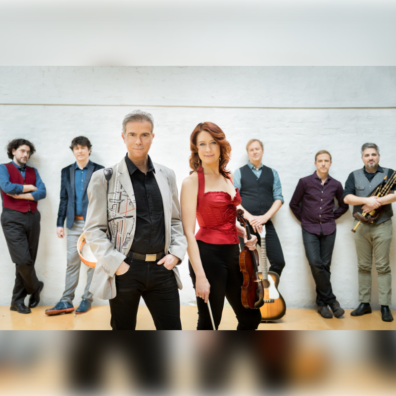 A group of seven musicians, including a violinist and a bagpiper, stand against a plain white wall.